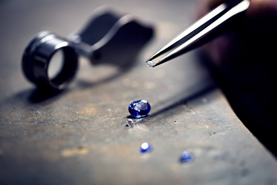 Tweezers placing a diamond beside an oval blue sapphire on a worn steel bench, with a jeweller's loupe